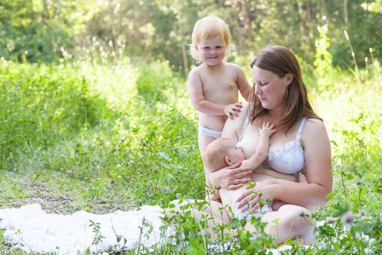 toddler stands with her mom as she breastfeeds her baby