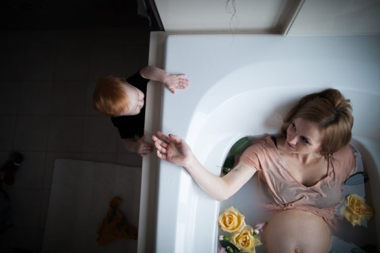 toddler peeks over edge of tub at his mama during her maternity pictures