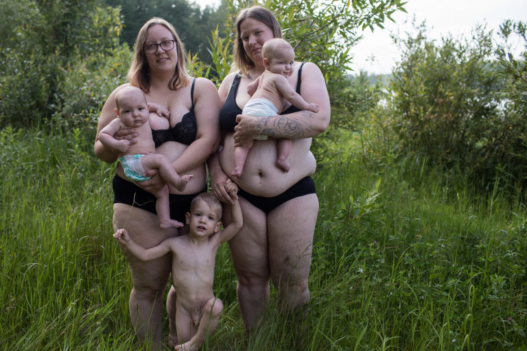 two moms stand holding their twins with their toddler standing in front of them